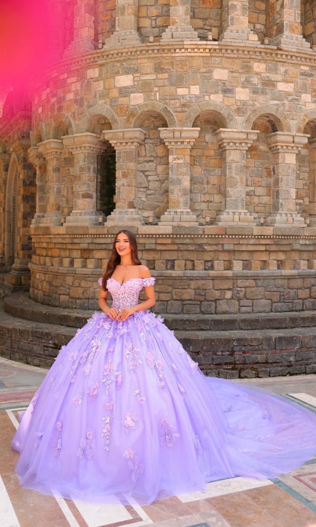 Wearing the Amarra 54231 3D Floral Quinceañera Dress, a woman in a lavender ball gown stands before a stone building with arched windows and columns.